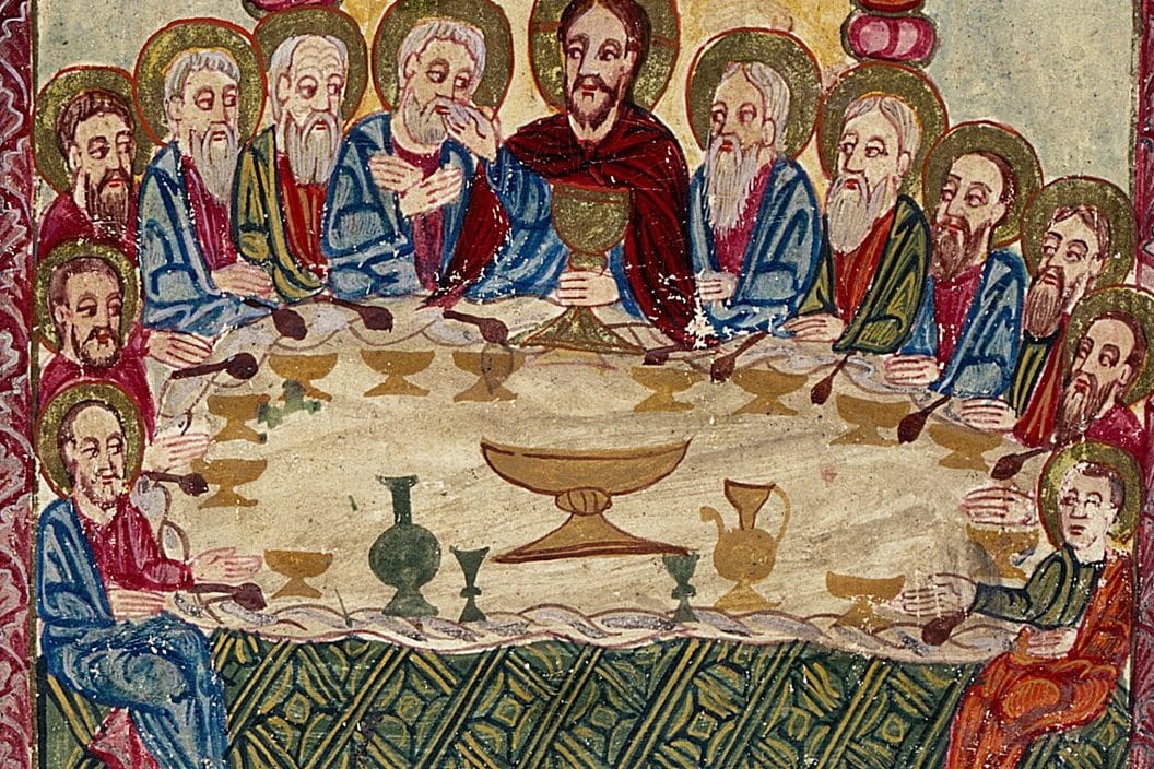Christ, in a red robe, sits at the center of a long table holding a golden chalice, surrounded by haloed apostles, with cups, a jug, and a wide bowl set on the white cloth.