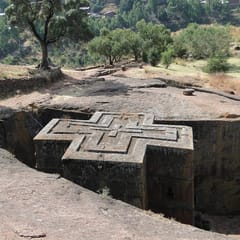 A church carved down into solid red rock in the shape of a cross, its flat roof marked with nested crosses, stands at the bottom of a deep trench cut into a hillside dotted with trees.