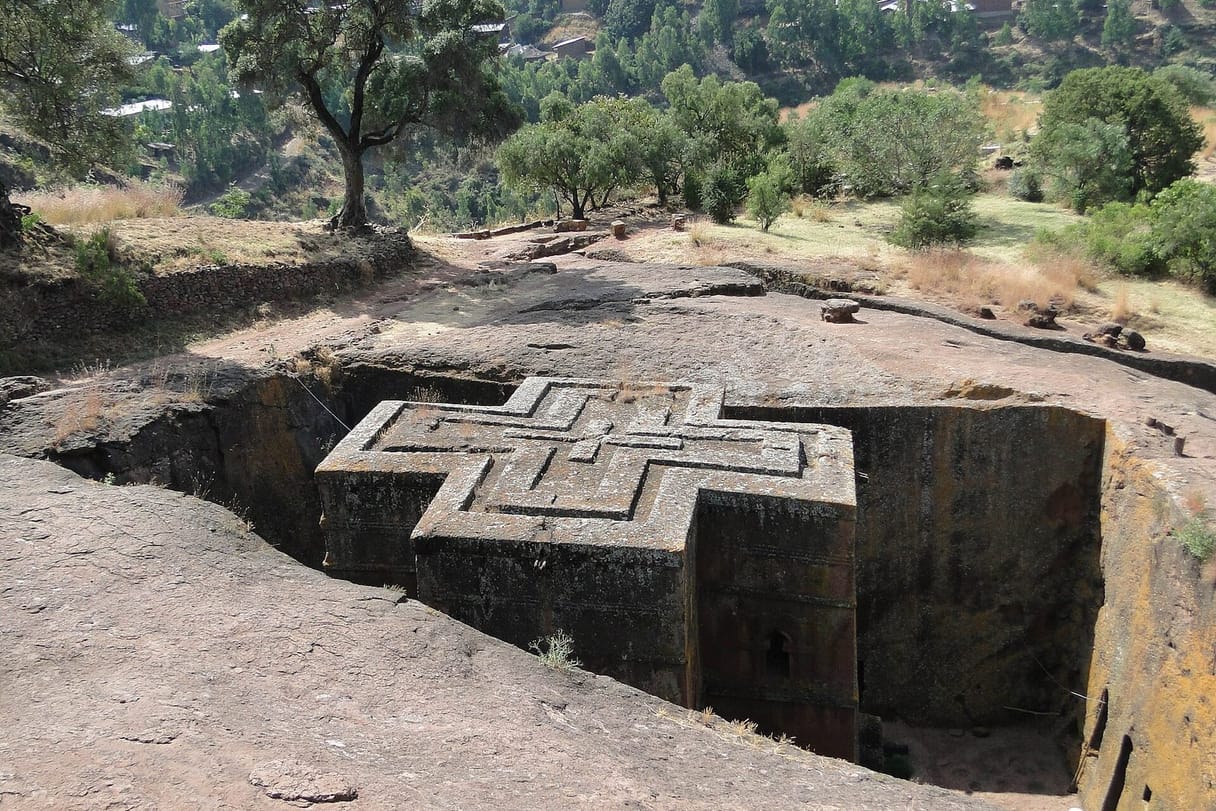 A church carved down into solid red rock in the shape of a cross, its flat roof marked with nested crosses, stands at the bottom of a deep trench cut into a hillside dotted with trees.