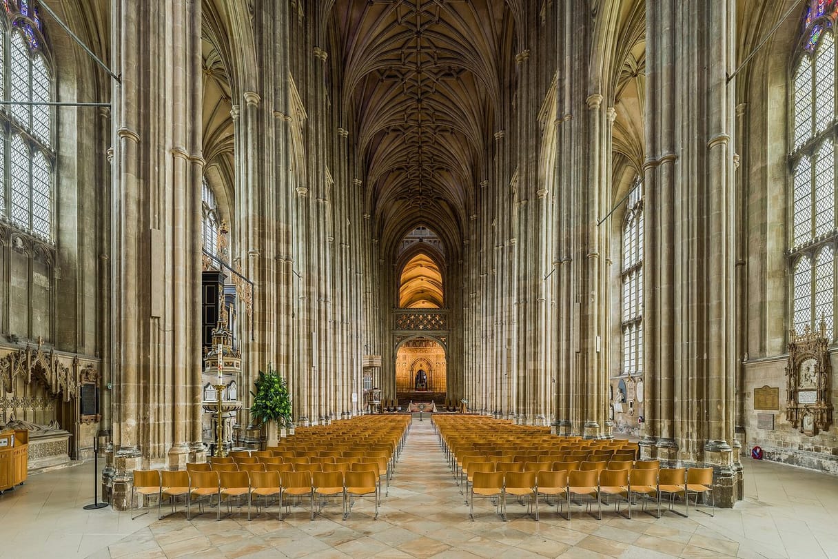 A long Gothic nave of tall clustered columns and fan-like vaulting leads the eye toward a lit stone screen at the far end.