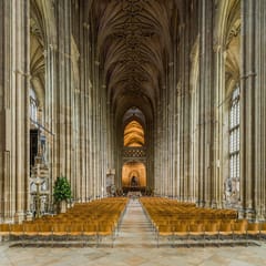 A long Gothic nave of tall clustered columns and fan-like vaulting leads the eye toward a lit stone screen at the far end.