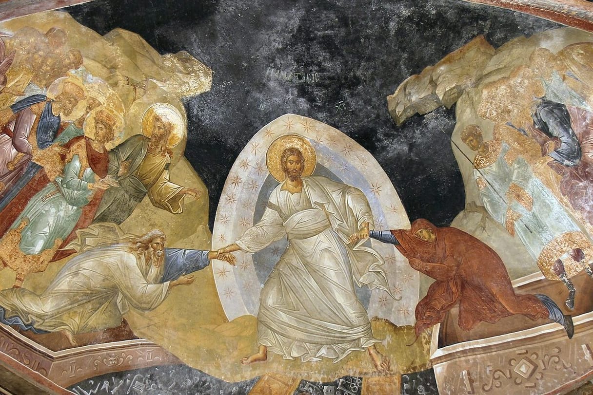 Christ in shining white robes stands on the broken gates of hell and pulls Adam and Eve up out of their tombs by the wrists.