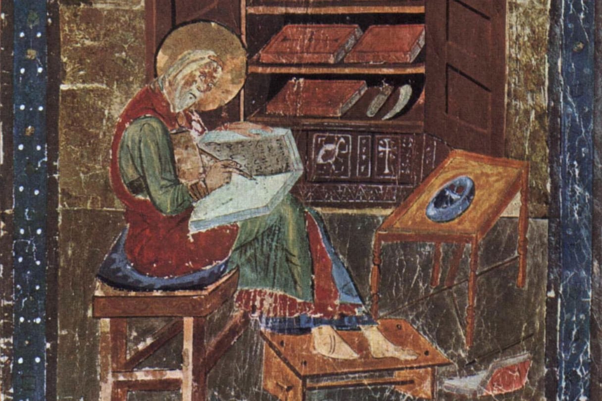 A haloed scribe sits on a stool writing in a book on his lap, in front of an open cupboard stacked with bound volumes.