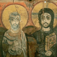 On a cracked wooden panel, Christ, holding a jeweled Gospel book, rests his arm on the shoulder of a gray-bearded abbot who stands beside him; both have large golden halos and wide, steady eyes.