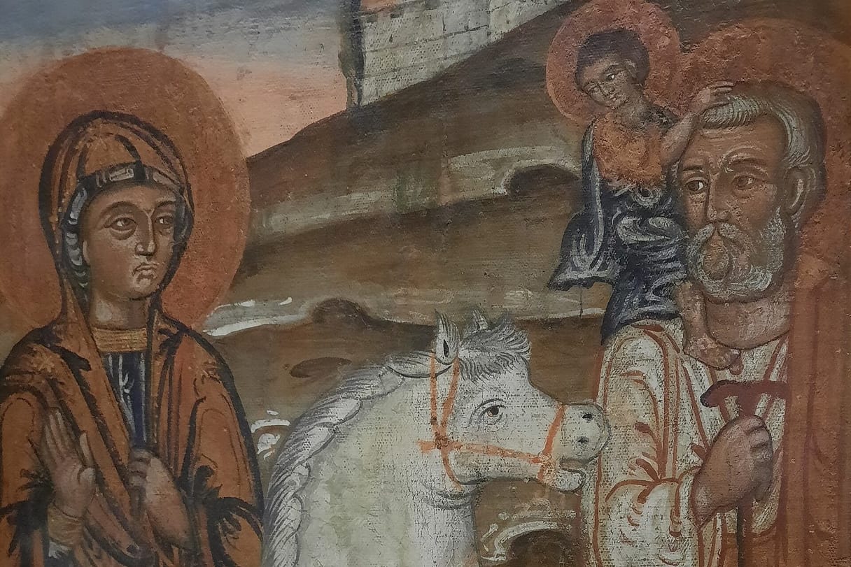 Mary, in a dark blue robe and brown mantle, rides a white mount while Joseph walks beside her carrying the young Jesus on his shoulders, below a walled city on a hill.