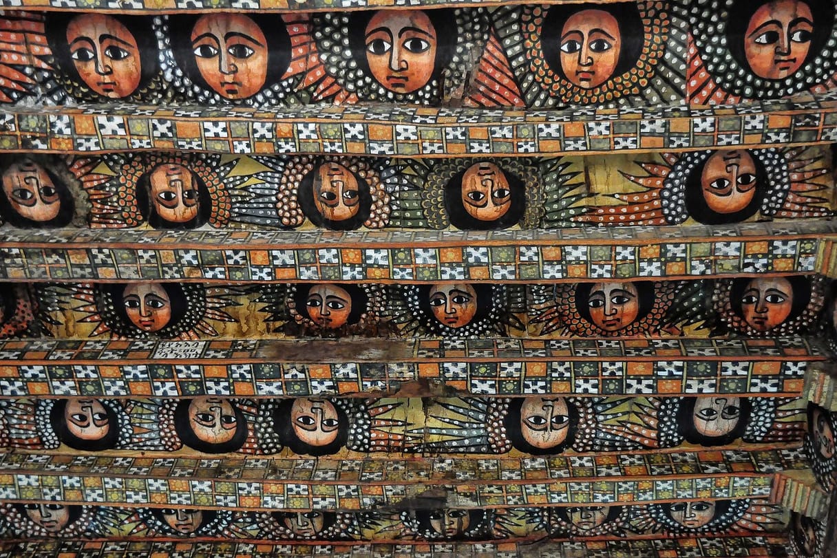 Rows of painted angel faces with large dark eyes and outspread wings fill the beams of a church ceiling, separated by bands of patterned crosses.
