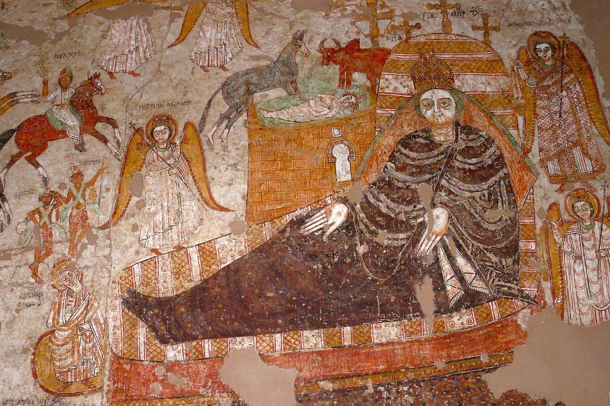 Mary, in a dark brown robe, reclines on a large patterned couch while the swaddled Christ Child lies in a manger above her, watched by an ox and a donkey; angels stand at the sides and the Magi ride in on horseback at the left.