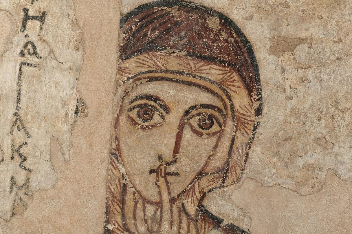 A wall painting of a woman's face framed by a dark veil, with large, wide-open eyes and one finger raised to her lips; a Greek inscription runs down the left side.