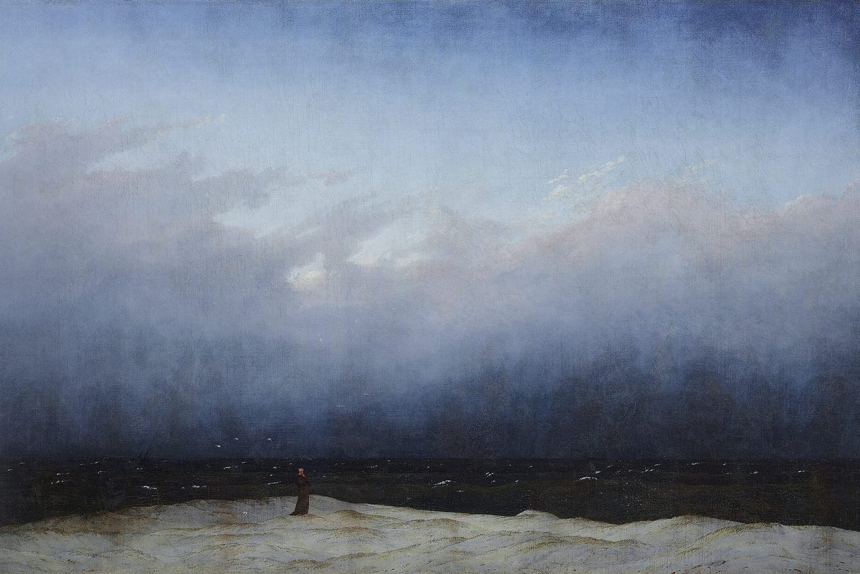 A tiny robed figure stands alone on a pale dune before a black sea, under an immense sky that darkens from misty blue to near night.