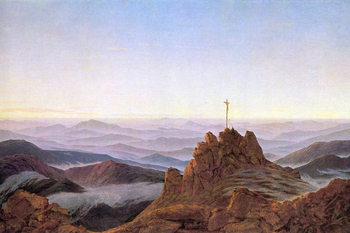 Morning light spreads over range after range of misty blue mountains, with a small cross standing on a rocky summit in the foreground.