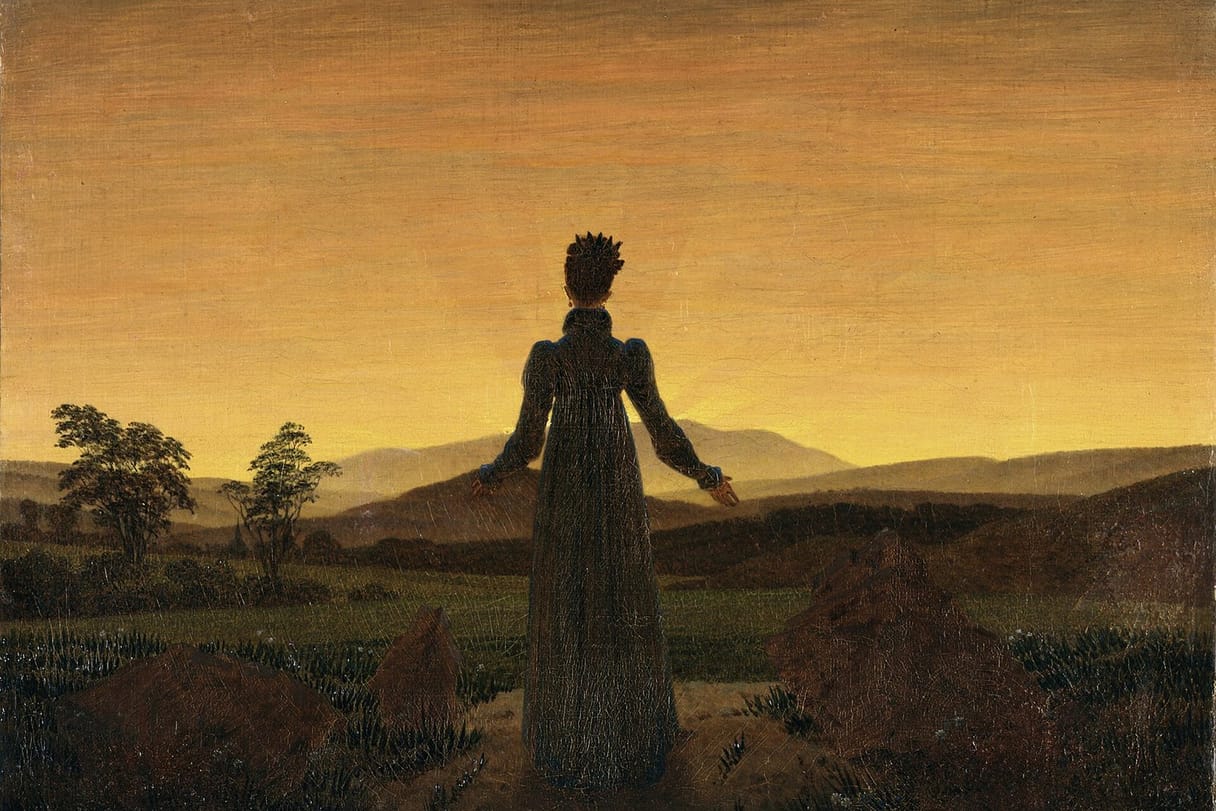 A woman seen from behind stands on a path with her arms opened toward a glowing golden sky over dark hills.