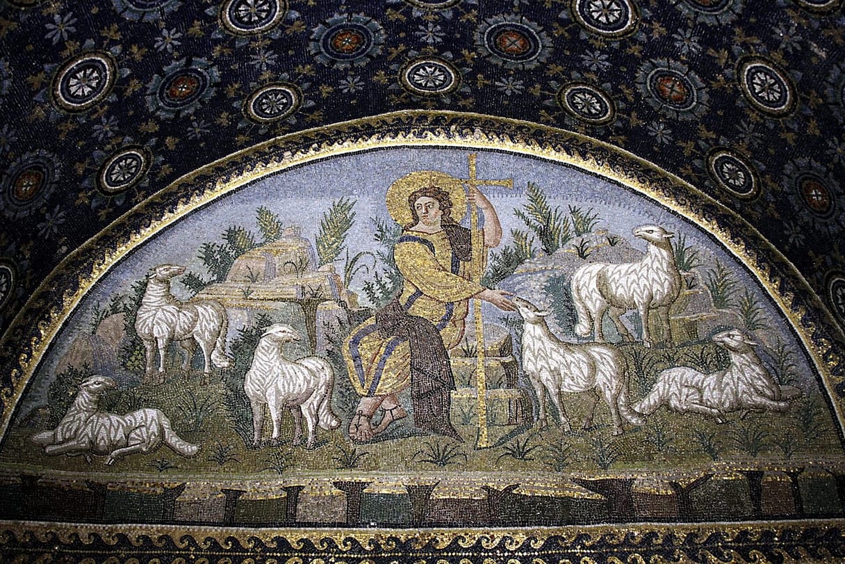 A youthful Christ in golden robes sits among white sheep on a rocky meadow, holding a tall cross, in a lunette of glittering mosaic.