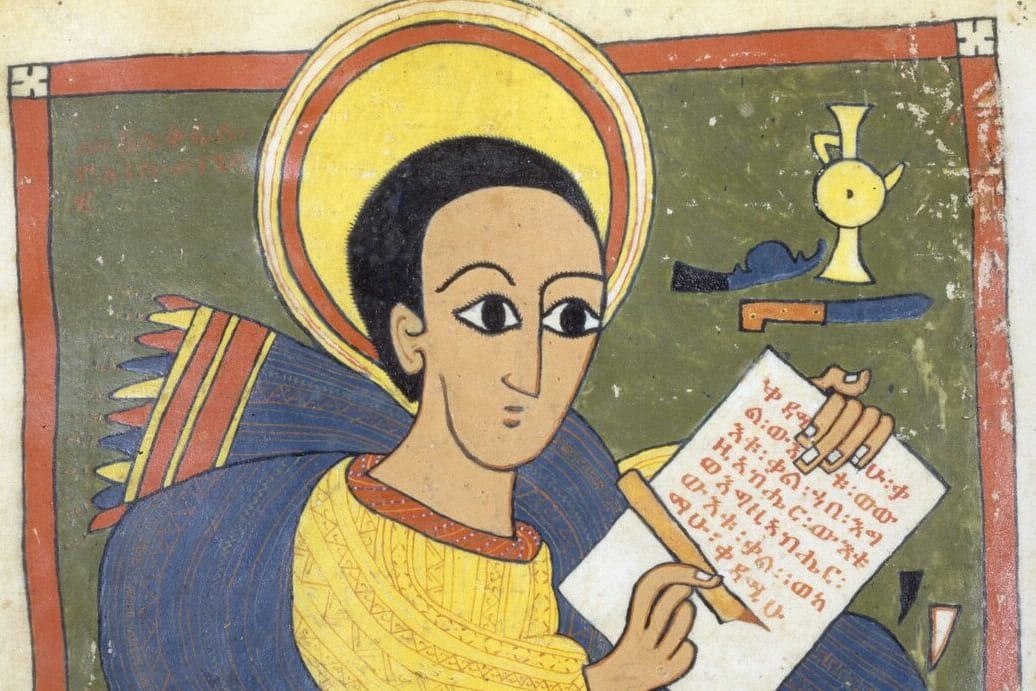 A young evangelist with a large golden halo and wide dark eyes writes lines of Ge’ez script on an open scroll, wrapped in a blue mantle over a yellow tunic, against a green ground.