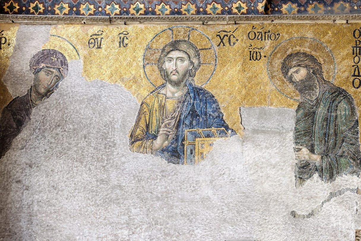 A gold mosaic of Christ holding a book and raising his hand in blessing, between the Virgin Mary and John the Baptist, who turn toward him; the lower half of the mosaic has been lost to bare plaster.