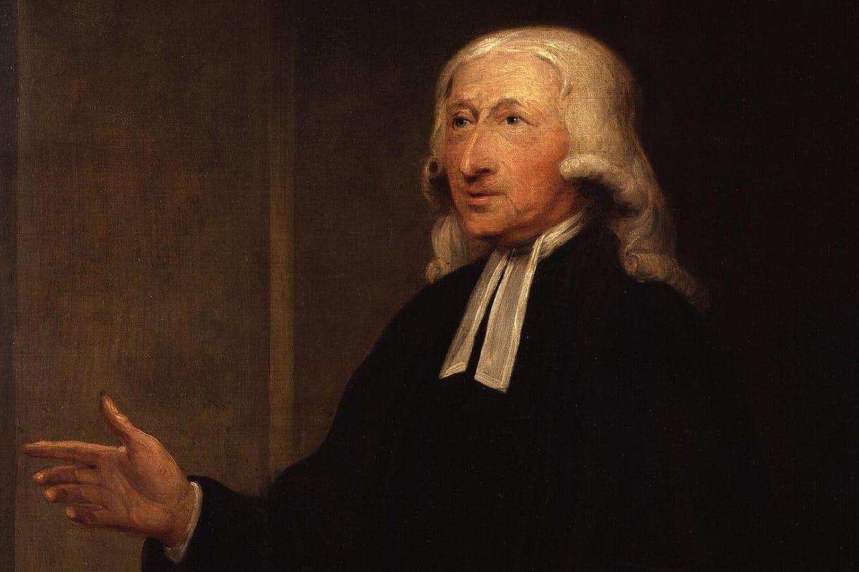An elderly clergyman with long white hair, a black gown, and white preaching bands looks to one side and gestures with an open hand as he speaks.