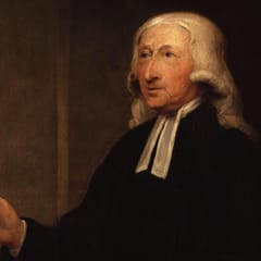 An elderly clergyman with long white hair, a black gown, and white preaching bands looks to one side and gestures with an open hand as he speaks.