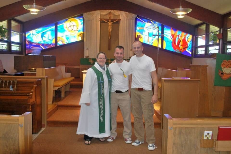 A smiling woman pastor in a white robe and green stole stands with two grooms in front of the altar and stained-glass windows of a Lutheran church.