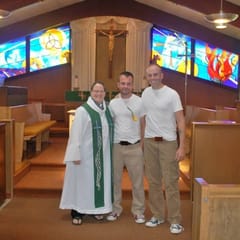 A smiling woman pastor in a white robe and green stole stands with two grooms in front of the altar and stained-glass windows of a Lutheran church.