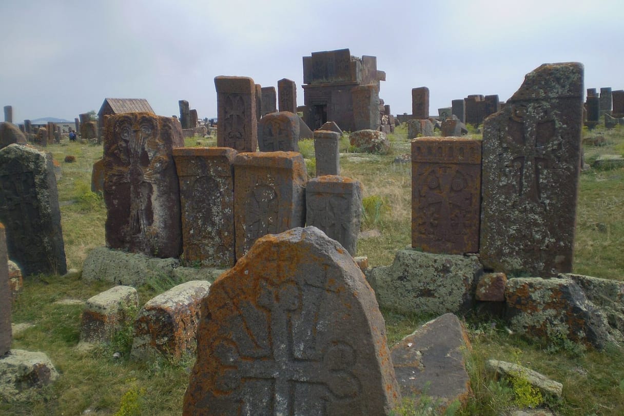 Weathered stone grave markers carved with crosses stand among long grass, the nearest a rounded slab with a cross in relief, under a pale sky.