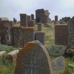Weathered stone grave markers carved with crosses stand among long grass, the nearest a rounded slab with a cross in relief, under a pale sky.