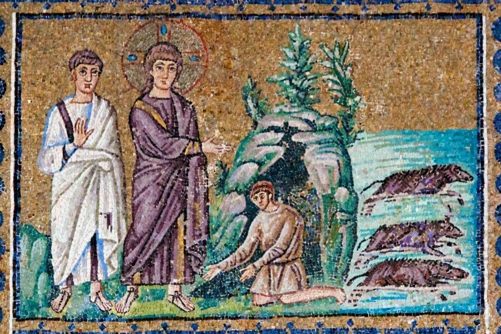 On a gold background, Jesus in a purple robe stretches out his hand toward a man kneeling at the mouth of a rocky tomb, while a disciple in white looks on and three pigs plunge into the sea.