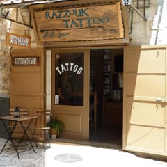 The open wooden doors of the Razzouk Tattoo shop, under a carved sign, on a sunny stone street in Jerusalem, with a small café table outside.