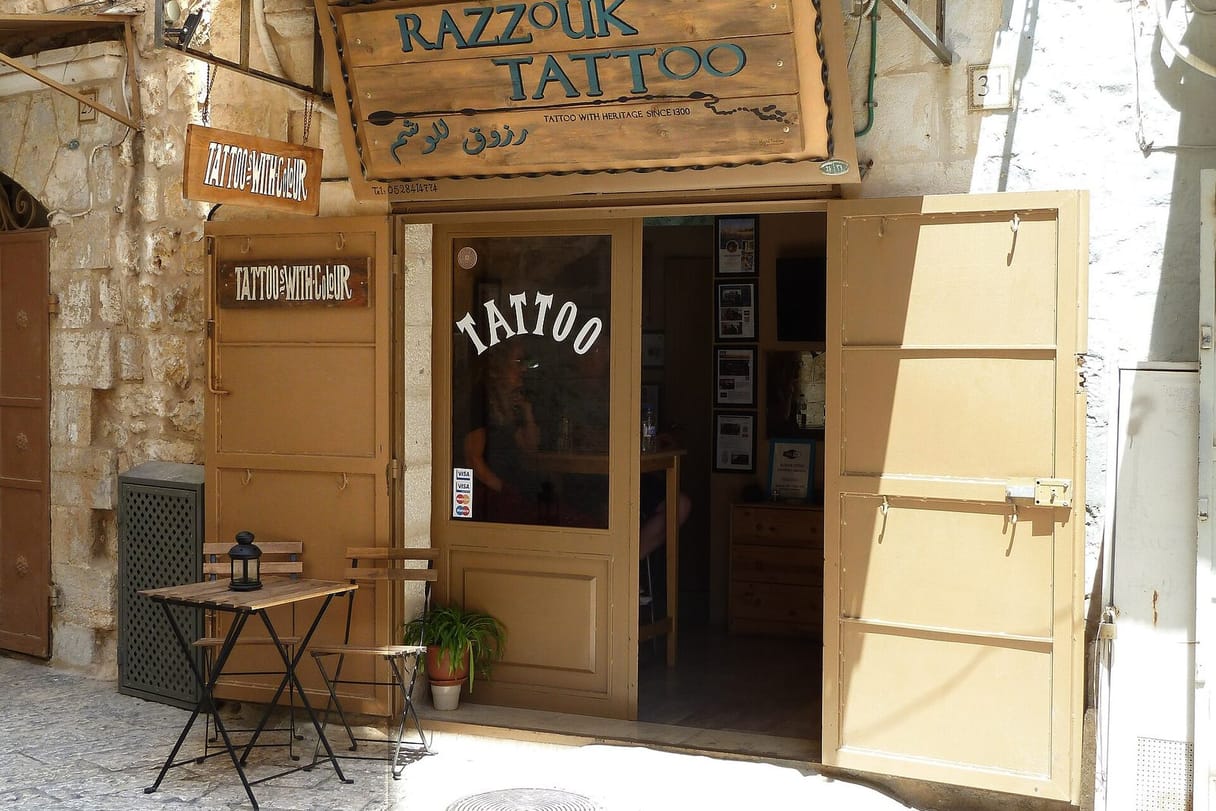 The open wooden doors of the Razzouk Tattoo shop, under a carved sign, on a sunny stone street in Jerusalem, with a small café table outside.