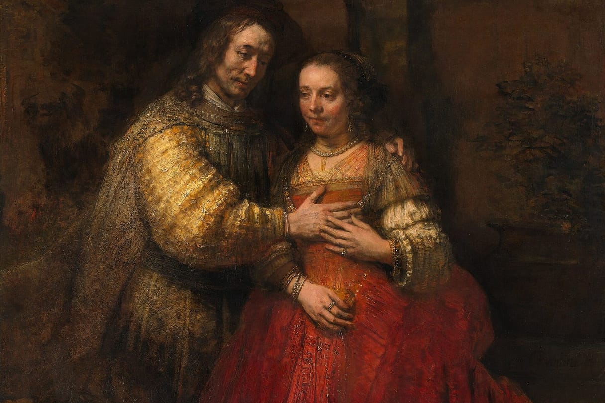 A husband in a golden sleeve gently rests his hand on his wife, who lays her own hand over his, both lost in thought.