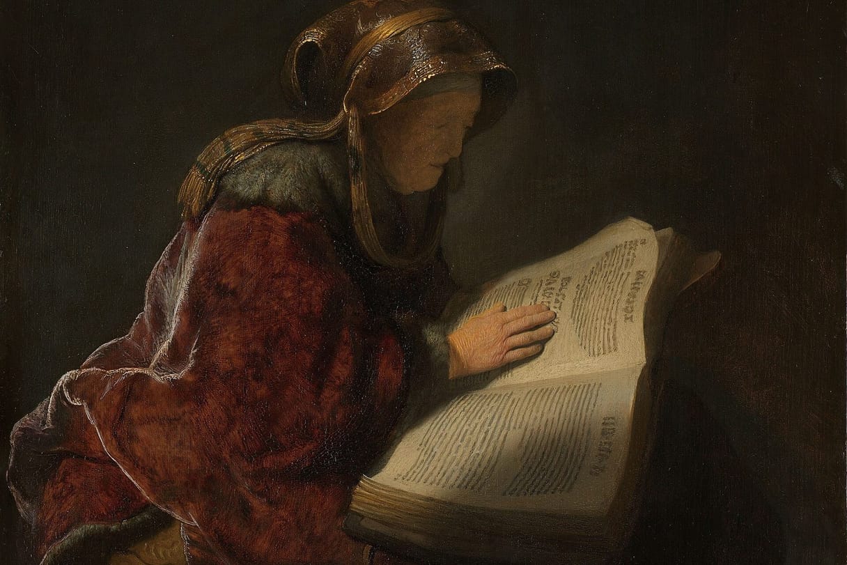 An old woman in a fur-trimmed red cloak and a gold headscarf reads a large open book, her hand resting on the page.