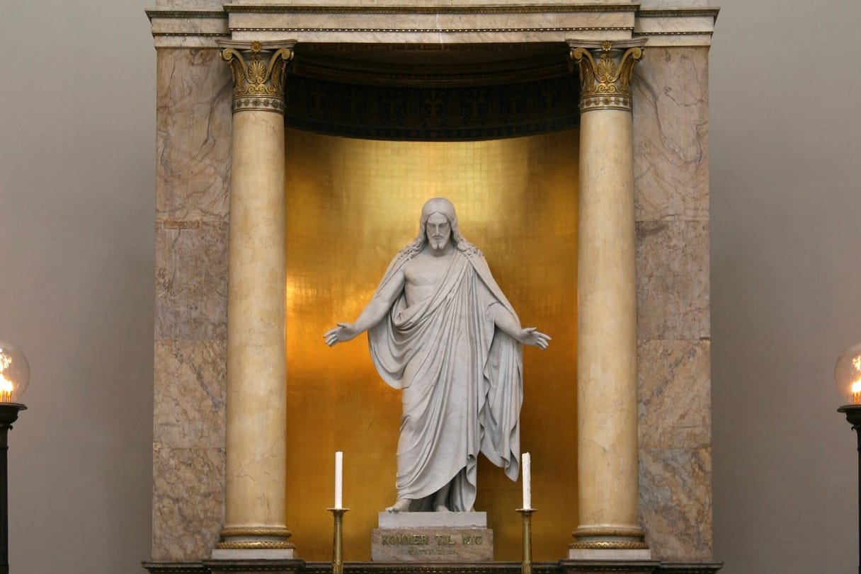 A white marble statue of Christ with open arms stands in a gilded niche between two marble columns above a church altar with candles.