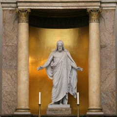 A white marble statue of Christ with open arms stands in a gilded niche between two marble columns above a church altar with candles.