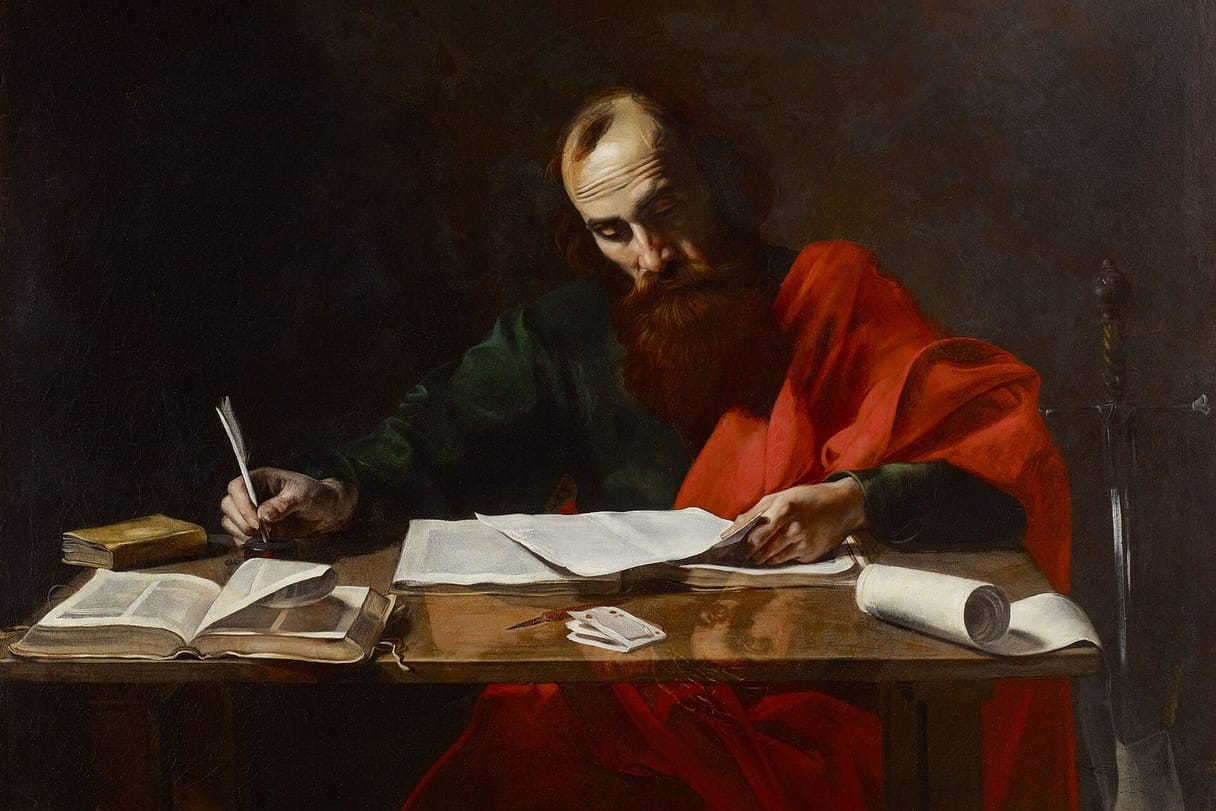 The apostle Paul, in a red cloak, sits at a table covered with books and papers, writing a letter with a quill.