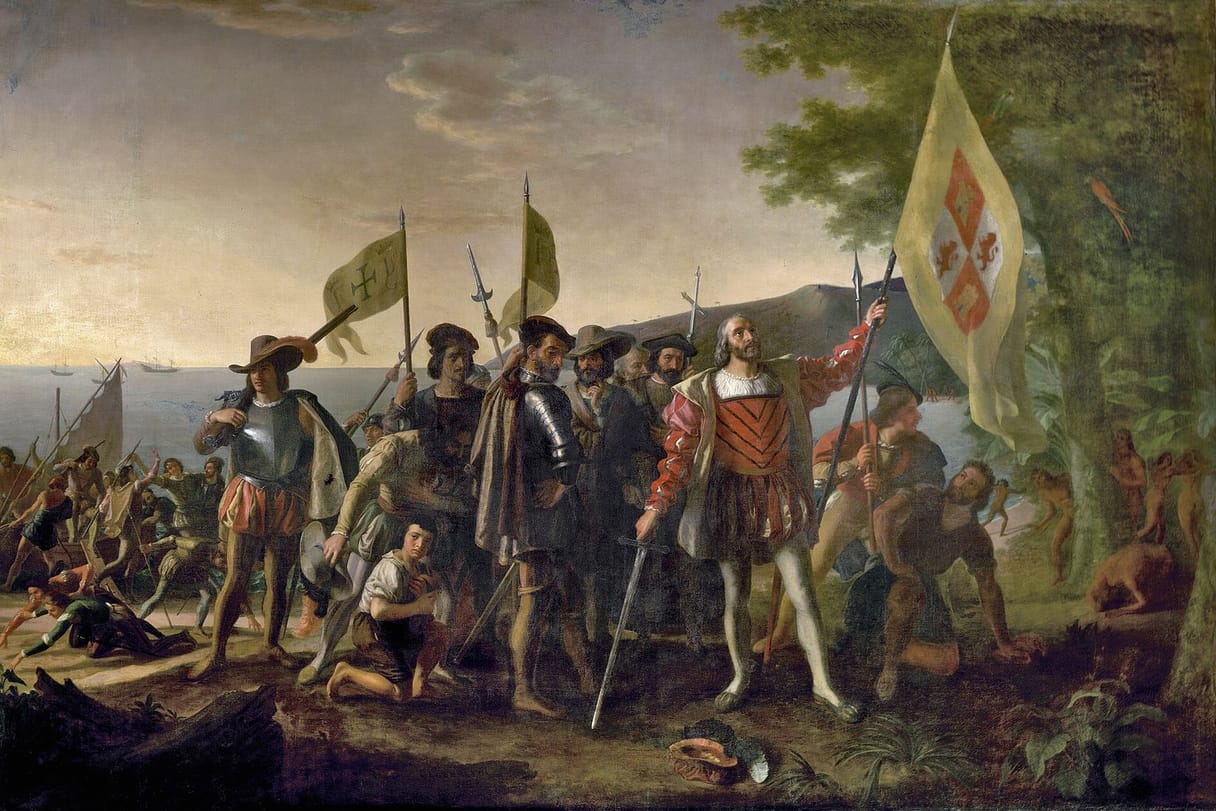 On a tropical shore, Columbus stands bareheaded holding the royal banner of Spain upright, a sword in his other hand, among armored officers and kneeling sailors, while Indigenous people watch from the trees at the right.