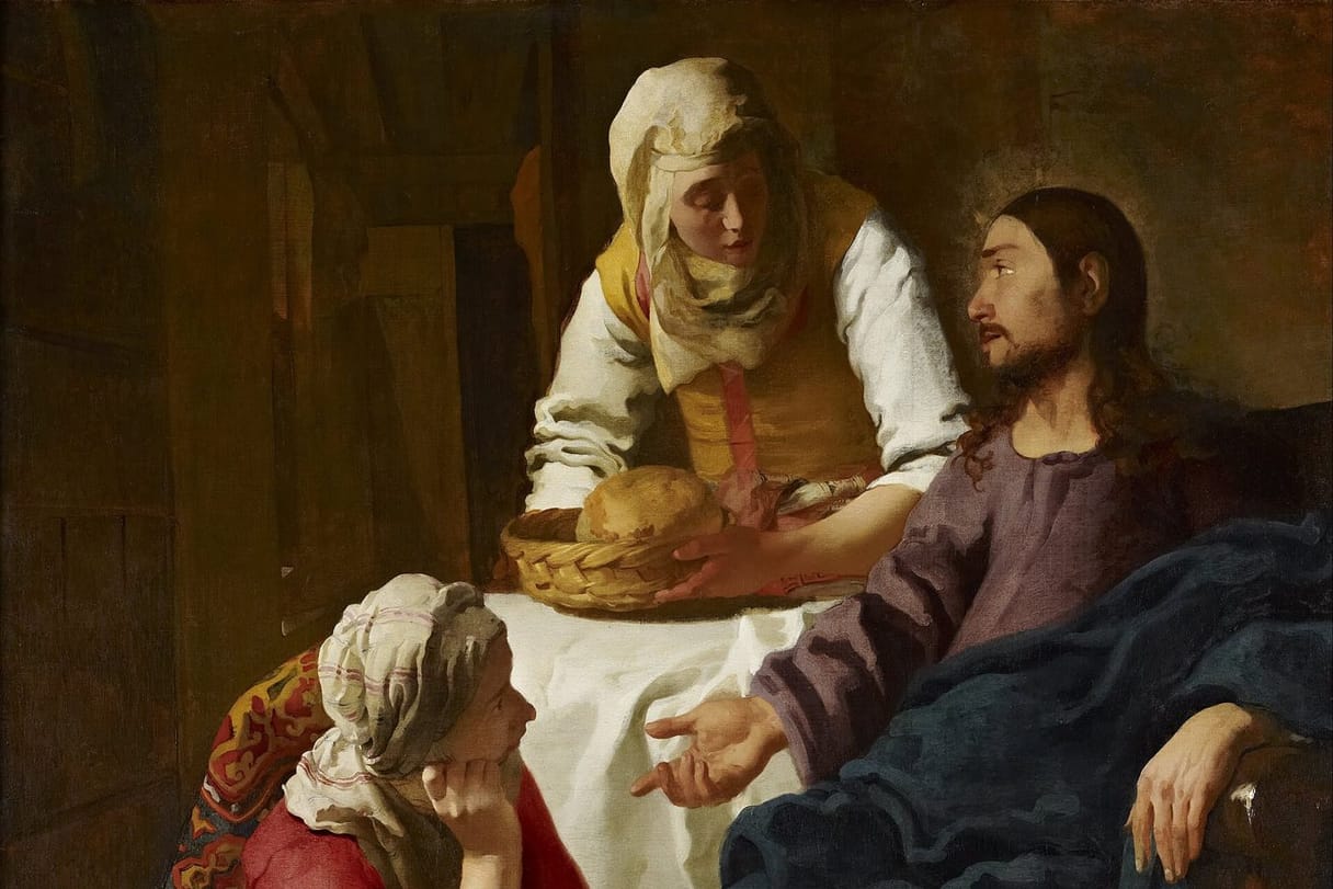 Jesus sits in a chair, gesturing toward a young woman seated at his feet, while her sister leans over the table holding a basket of bread.