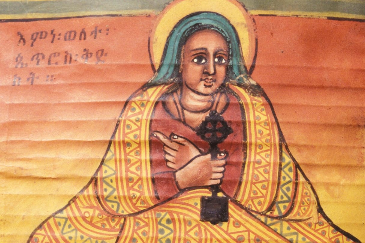 A haloed woman in a blue headscarf and a yellow patterned mantle sits facing us, holding a dark hand cross against her chest, beside her name written in Ge’ez.