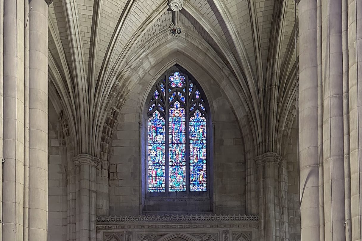Pale stone columns and ribbed Gothic vaults frame a tall stained-glass window in a quiet bay of the cathedral.