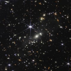 Thousands of galaxies, some stretched into thin arcs, glow gold and white against the black of deep space.