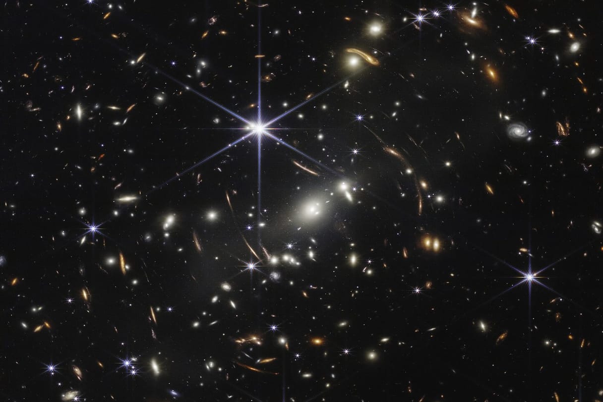Thousands of galaxies, some stretched into thin arcs, glow gold and white against the black of deep space.